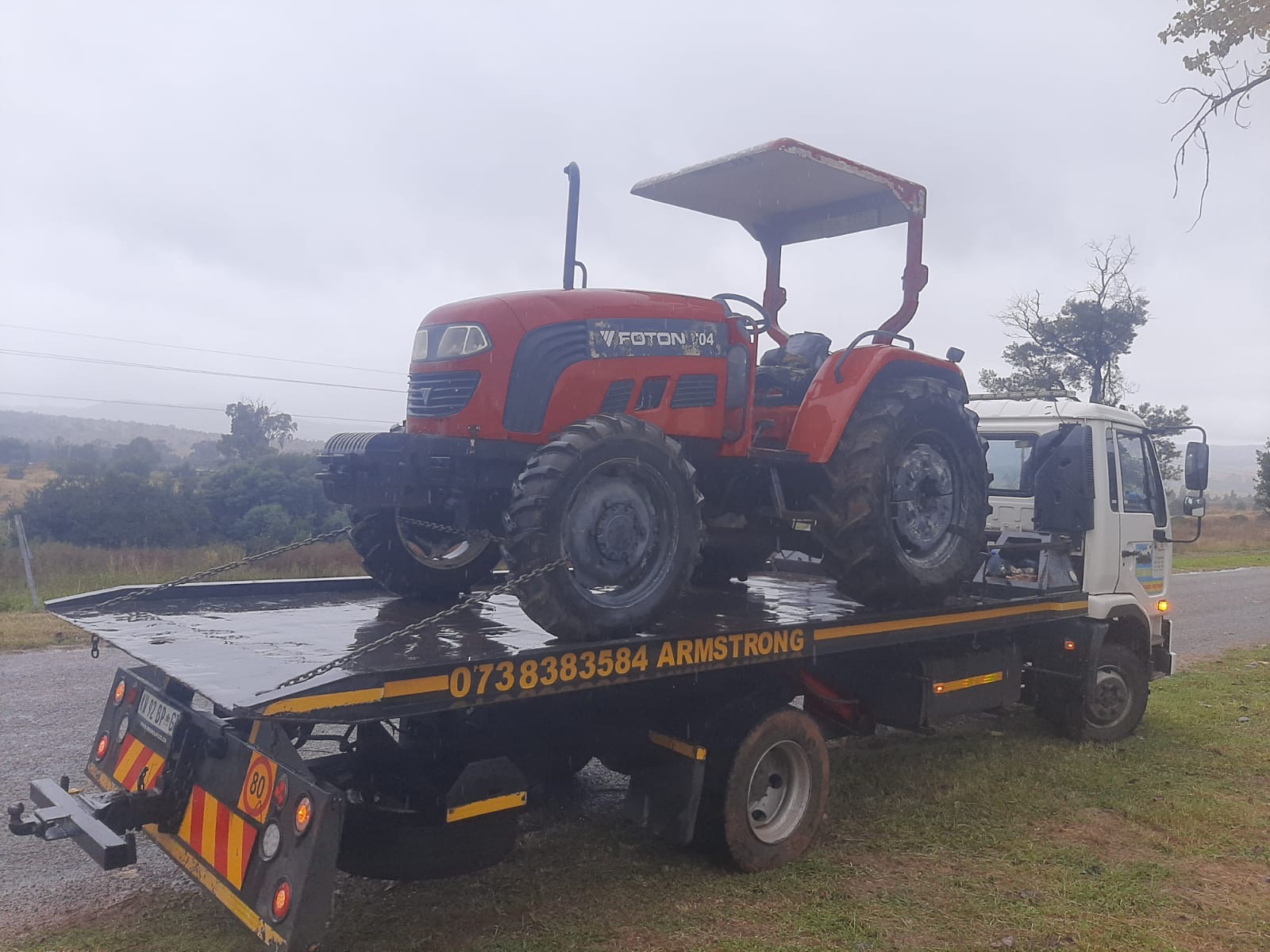Red Foton tractor chained down on the Armstrong Towing flatbed in the rain
